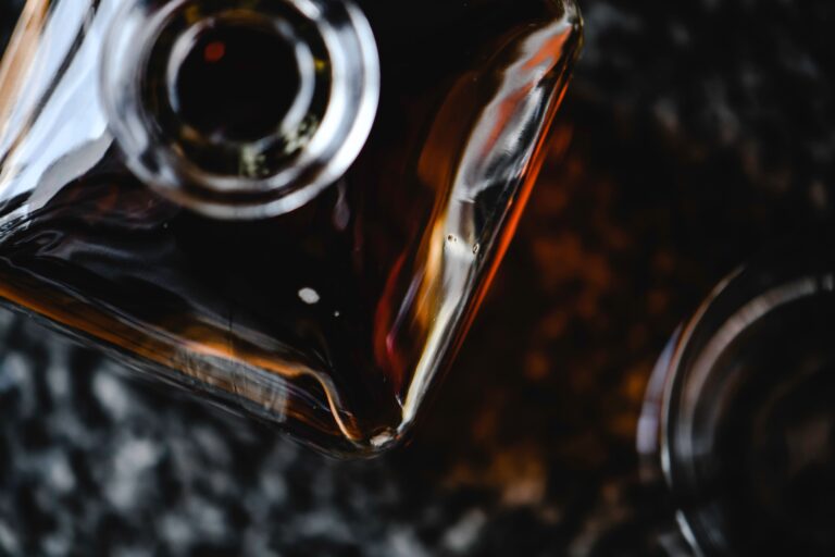 Zbliżenie na szklaną butelkę z grubym dnem, wypełnioną ciemną bursztynową whisky, sfotografowaną z góry w kontrastowym oświetleniu na ciemnym tle.